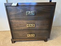 Front angle view of dark wood three drawer dresser showing Asian motif brass or gold-tone rectangular drawer handles, solid wood with visible grain.