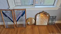 Two wooden framed stained glass panels, one round mirror, one rectangular mirror with ornate frame, and one metal framed stained glass panel with crack all standing on the floor near window.