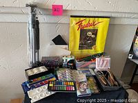 Overall lot showing a foldable tripod easel in plastic wrap, Fredrix artist paper, multiple paint sets in pans and tubes, boxes of pencils and brushes.