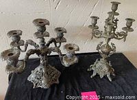 Photo showing both antique 5-arm candelabras side by side on a black cloth surface with visible ornate details and tarnish.