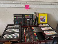 Full view of the open wooden artist kit case showing multiple compartments with acrylic paint tubes, colored pencils, pastels, drawing pencils, eraser, sharpener, and watercolor palette.