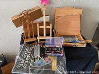 Overall lot showing three small wooden easels, wooden artist box with brushes, colored pencils, paints, and paper palette on a black fabric surface.