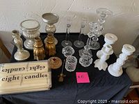 Assorted candle holders and two boxes of white taper candles on a black cloth-covered table, displaying the variety of sizes and styles.