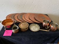 Photo showing all items in the lot arranged on a black cloth: 8 copper plates, 2 copper mugs, small frying pan, colander, pitcher, and lidded pots.