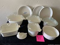 Full view of CorningWare set including three casserole dishes with glass lids showing the blue cornflower design, three matching plates, white ceramic hen container, and round fluted ceramic dish.