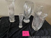 Full view of the five assorted crystal vases of various shapes and sizes placed on black fabric against a textured wall.