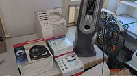 Photo showing the Honeywell Enviracaire air purifier and Lasko tower fan with boxes beneath them in a room.