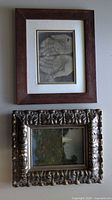 Two framed artworks shown together; one drawing of hands in dark wood simple frame and one landscape painting in ornate wood frame