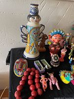 Photo showing an assortment including a tall ceramic vase with a female face on the neck, a red beaded necklace, colorful masks, and small figurines.