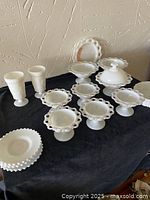 Full view of assorted milk glass tableware on black cloth background including hobnail edge pedestal plates, goblets, and pedestal compote dish.