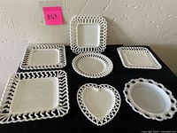 Front view of seven milk glass plates arranged on black fabric, showing various square, round, and heart shapes with looped rims