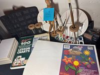 Overview of ceramic paint plates, wooden figure, glass and cork brush holders, watercolor palettes, paint brushes, watercolor lesson charts, and canvas boards captured on table.