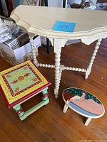 Front and overall view of off-white painted wooden table with two colorful small stools in front: one square with folk patterns, one oval with nude figure painting