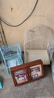 Photo showing two bird cages; one large white cage with curved top and one smaller blue cage, along with a small wooden bird cabinet with glass-front doors that have bird-themed artwork.