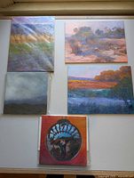 Five paintings displayed on floor: four landscapes two wrapped in plastic, one self-portrait painting with circular frame and orange background unwrapped