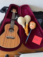 Ukulele inside red velvet-lined hard case with two white maracas and a decorative stick with a painted head.