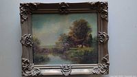 Full view of framed oil painting of a house in a landscape scene with water and trees.