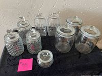 Full lot showing all nine glass jars with lids of various shapes and sizes on a black cloth background against a textured off-white wall.