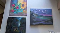 Photo showing three acrylic paintings, including the floral still life with roses and sunflower, a nighttime landscape with moon, and a waterfall landscape.