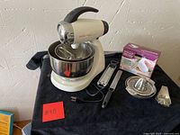 Wide shot of entire lot showing vintage Sunbeam Mixmaster stand mixer with attachments, Brentwood hand mixer box, kitchen grater, citrus juicer, and metal kitchen scale on black table cloth against textured wall.