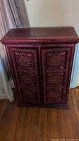 Front angled view of the wooden cabinet, showing dark red finish and gold painted patterns on the top and doors.