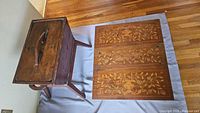 Wooden chest placed beside the wooden table with floral inlay panels folded out on the floor covering.