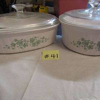 Two white CorningWare casserole dishes with green ivy leaf pattern and ribbed design, each with clear glass lids on beige cloth.