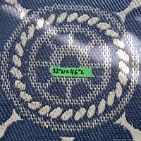 Close-up of blue outdoor rug portion showing central ship wheel pattern and rope border with size label 32"W x 46"L.