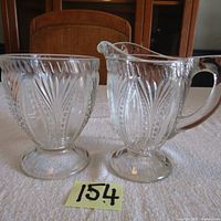 Two pressed glass items: a sugar bowl and a creamer pitcher, both with detailed pressed glass patterns and footed bases, displayed on a white fabric surface with a number 154 in front.
