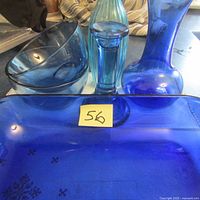 Photo showing two rounded blue glass bowls, one tall slender ribbed blue glass vase, one shorter smooth blue glass vase, one rectangular blue glass baking dish with measurement label, and one cylindrical blue glass candle holder.