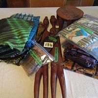Lot containing stacks of folded colorful African fabrics/textiles, pair of hand carved wooden figures lying flat, small wooden stool, and bags labeled 'real snake skin' and 'hand carved'.