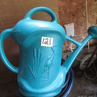 Blue plastic watering jug with molded design placed on the floor with hose nearby.