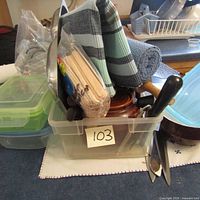 Plastic container filled with various kitchen towels, utensils, and wooden sticks bundled in plastic.