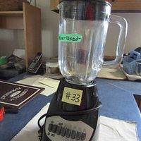 Black and Decker blender showing the clear plastic pitcher with a handle and a label 'Never Used'. The base is black and has a power cord wrapped around it.