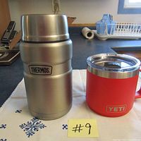 Photo of stainless steel Thermos jar with the Thermos logo and the red Yeti Rambler mug with handle and logo on a table