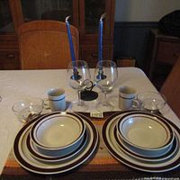 Full view of all tableware and accessories set for two on a white tablecloth including plates, bowls, mugs, glasses, placemats, and candle holder.