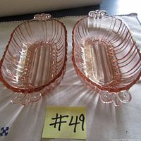 Two pink depression glass dishes side by side on white cloth with white background, showing ribbed pattern, oval center, and curled feet.