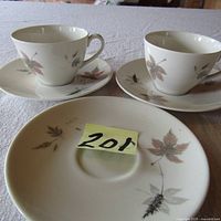 Two bone china cups with handles and three matching saucers featuring autumn leaf pattern in muted brown and gray colors, placed on a white textured cloth.