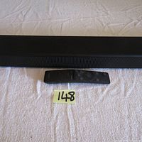Samsung sound bar with remote control on white fabric surface, labeled 148.
