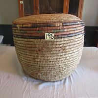 Full view of the very large woven wicker basket showing its rounded shape, multicolored bands near the top, and signs of wear. Basket is placed on a table against a cabinet background.