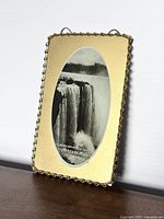 Frontal view of the antique oval black and white Niagara Falls Horseshoe Falls photograph mounted on decorative plate with twisted gold-tone border, placed upright against a wall on wooden surface.