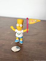 Front view of Bart Simpson Camp Krusty figure standing on a surface next to a coin for size comparison.