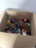 Top view of large cardboard box filled with various small toy cars and vehicles in different colors and styles.