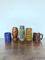 Five tiki mugs displayed in a row showing varied designs and colors.