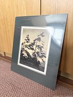 Full view of the sealed matted print leaning against a wood wall, showing the grey mat and visible print inside.