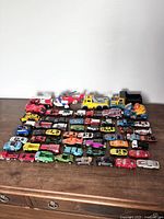 Front angled view of entire toy car and vehicle collection laid out on wooden surface, showing variety of vehicle types and colors, including toy trucks, cars and a plane