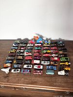 Top-down view showing 49 toy cars arranged in rows on a wooden surface. Cars include various colors and types such as racing cars, trucks, and special vehicles like a white space shuttle.