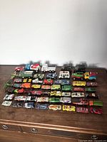Wide shot of 37 vintage toy cars arranged in rows on a wooden surface including trucks, emergency vehicles, sports cars, and sedans in multiple colors.