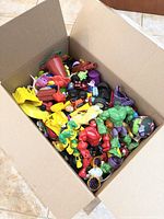 Open cardboard box with various small plastic toys including action figures, toy vehicles, and accessories