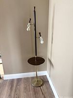 Full view of the floor lamp showing two light bulbs and a circular wooden table platform mounted on a brass pole with black accents.